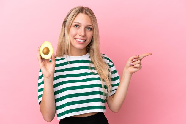 Young caucasian woman holding an avocado isolated on pink background pointing finger to the side