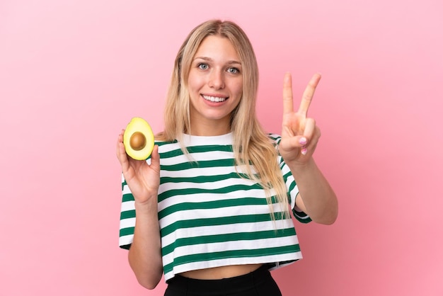 Young caucasian woman holding an avocado isolated on pink background smiling and showing victory sign