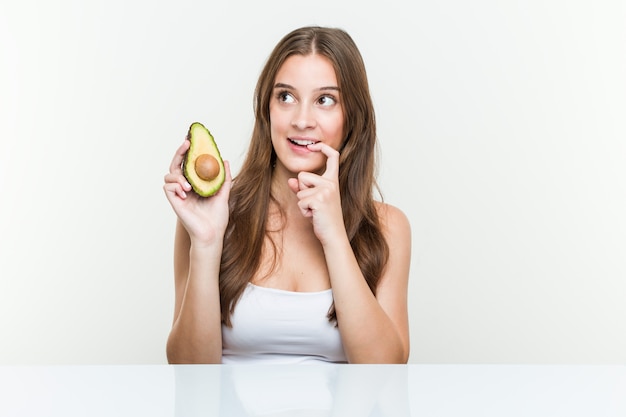 La giovane donna caucasica che tiene un avocado si è distesa pensando a qualcosa che osserva.