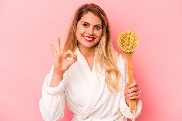 Young caucasian woman holding back scratcher isolated on pink background cheerful and confident showing ok gesture