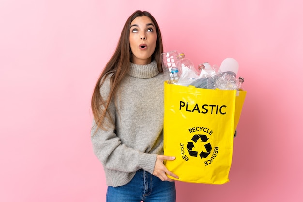 Young caucasian woman holding a bag full of plastic bottles to recycle isolated on pink background looking up and with surprised expression