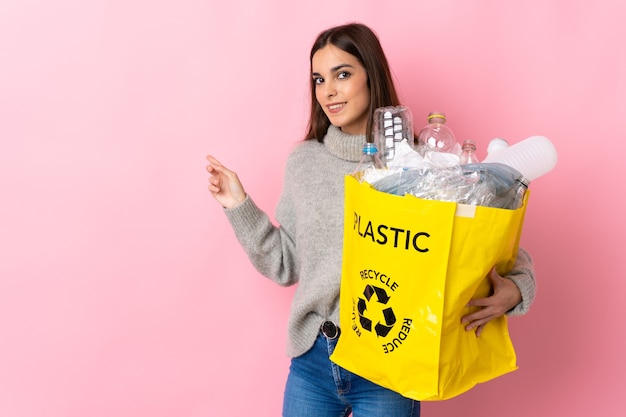 Foto giovane donna caucasica che tiene un sacchetto pieno di bottiglie di plastica da riciclare isolato su rosa che punta indietro