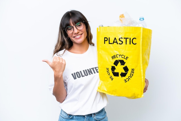 Young caucasian woman holding a bag full of plastic bottles to recycle isolated on white background pointing to the side to present a product