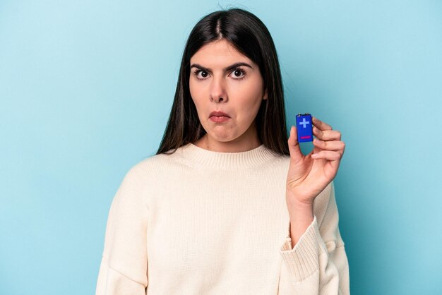 Young caucasian woman holding a batterie isolated on blue background shrugs shoulders and open eyes confused