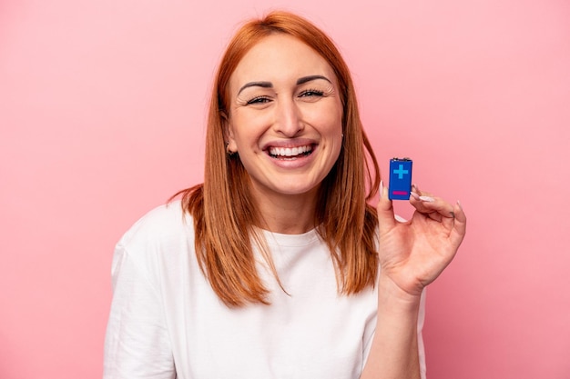 Young caucasian woman holding a batteries isolated on pink background laughing and having fun.