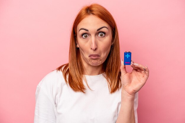 Young caucasian woman holding a batteries isolated on pink background shrugs shoulders and open eyes confused
