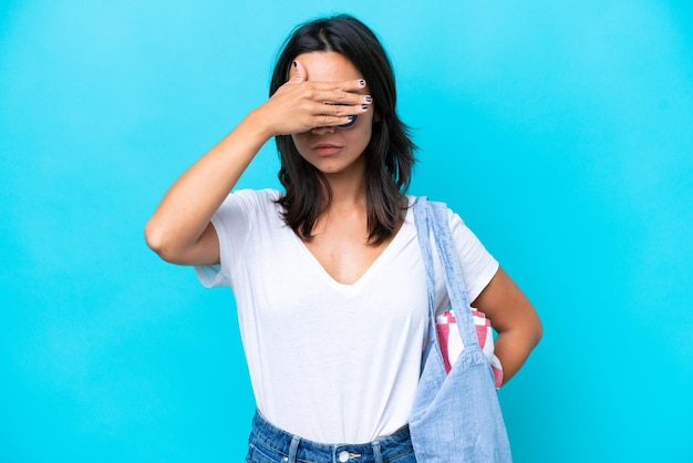 Young caucasian woman holding a beach bag isolated on blue background covering eyes by hands Do not want to see something