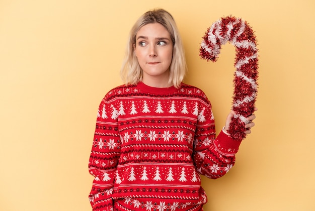 写真 黄色の背景に分離された大きなクリスマスの棒を持っている若い白人女性は混乱し、疑わしく、不安を感じています。
