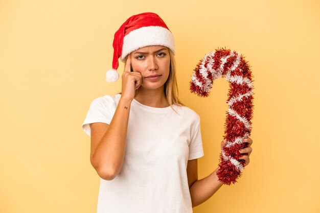 指で寺院を指して、黄色の背景に分離された大きなクリスマスの棒を保持している若い白人女性は、タスクに焦点を当てて考えています。