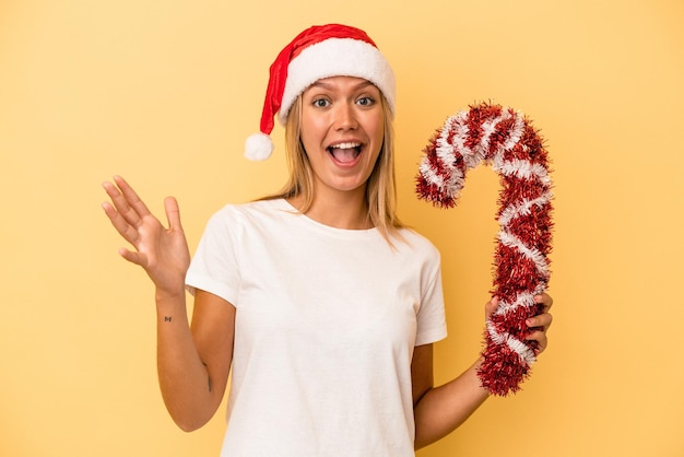 Young caucasian woman holding big christmas stick isolated on yellow background receiving a pleasant surprise, excited and raising hands.