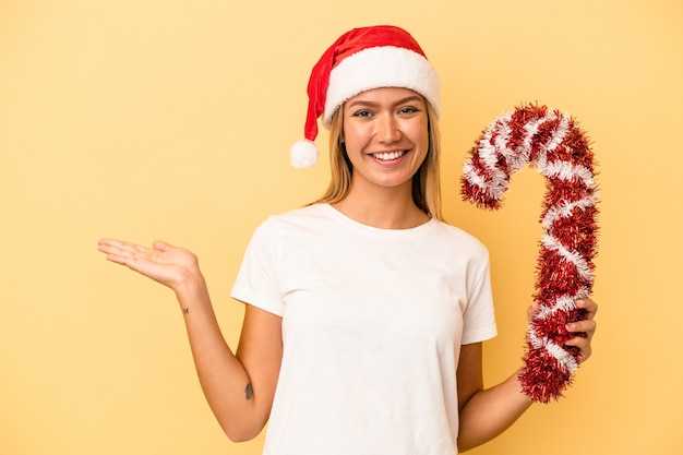 Photo young caucasian woman holding big christmas stick isolated on yellow background showing a copy space on a palm and holding another hand on waist.