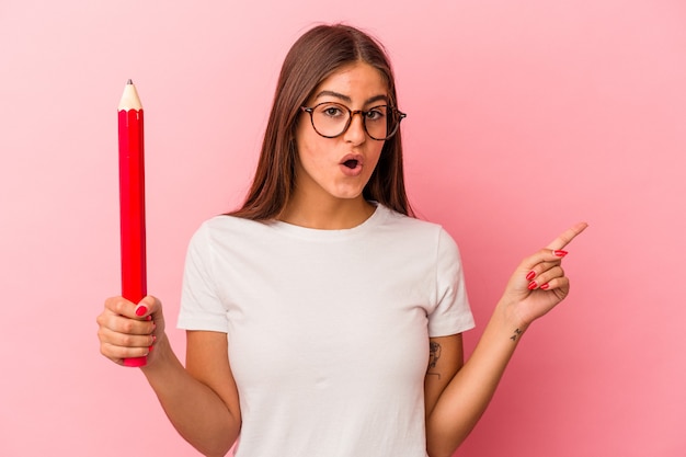 측면을 가리키는 분홍색 배경에 고립 된 큰 연필을 들고 젊은 백인 여자