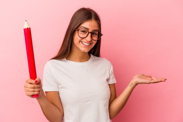 Young caucasian woman holding a big pencil isolated on pink background showing a copy space on a palm and holding another hand on waist.