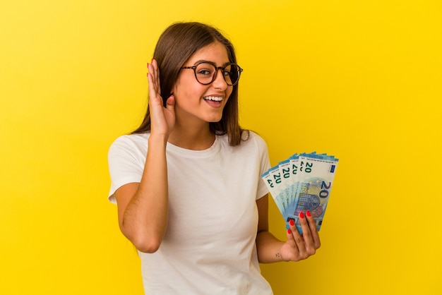 Young caucasian woman holding bills isolated on yellow background trying to listening a gossip.