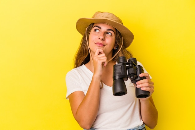 Young caucasian woman holding binoculars isolated on yellow background  looking sideways with doubtful and skeptical expression.