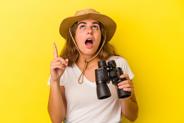 Young caucasian woman holding binoculars isolated on yellow background  pointing upside with opened mouth.