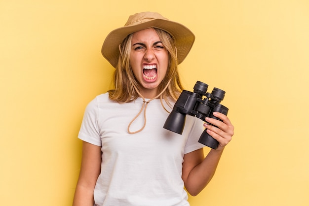 Young caucasian woman holding binoculars isolated on yellow background  screaming very angry and aggressive.