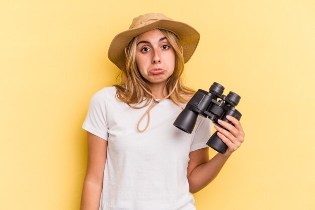 Young caucasian woman holding binoculars isolated on yellow background  shrugs shoulders and open eyes confused.