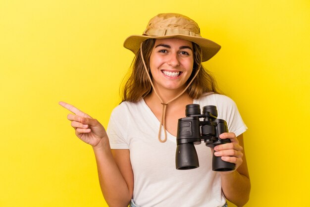 Foto giovane donna caucasica che tiene il binocolo isolato su sfondo giallo sorridente e puntato da parte, mostrando qualcosa in uno spazio vuoto.