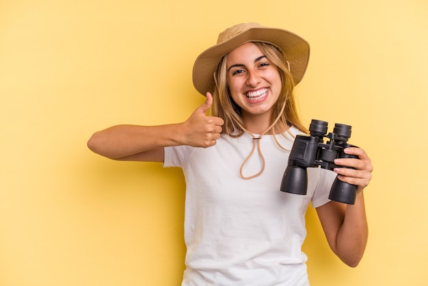 Young caucasian woman holding binoculars isolated on yellow background  smiling and raising thumb up