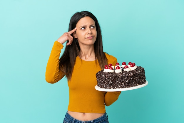 疑問と思考を持って青い背景で隔離のバースデーケーキを保持している若い白人女性