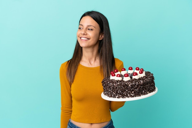 Young caucasian woman holding birthday cake isolated on blue background looking to the side and smiling