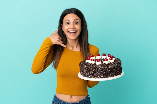 Young caucasian woman holding birthday cake isolated on blue background making phone gesture. Call me back sign