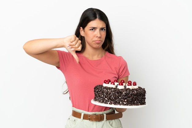 Young caucasian woman holding birthday cake isolated on white background showing thumb down with negative expression