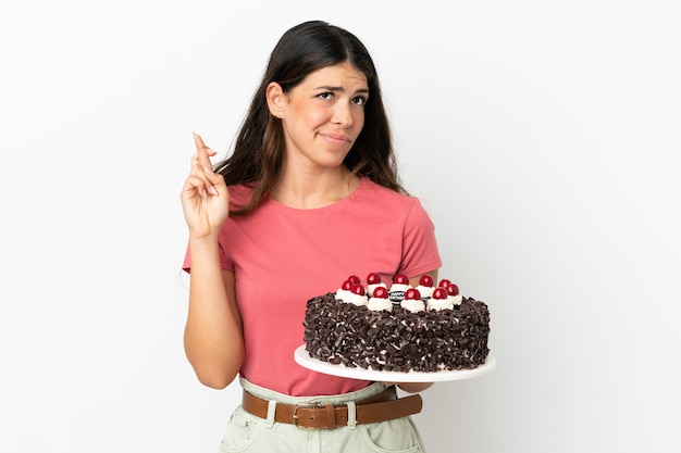 Young caucasian woman holding birthday cake isolated on white background with fingers crossing and wishing the best