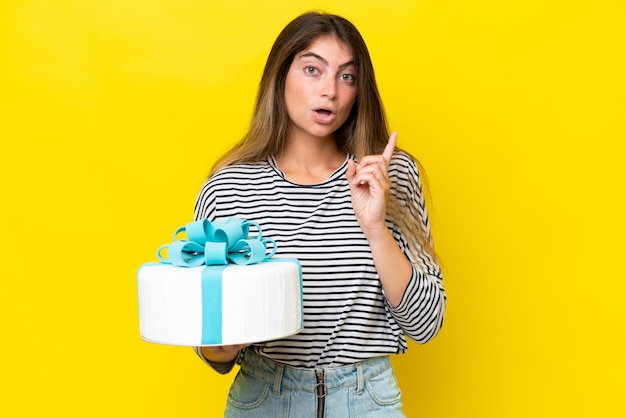 Young caucasian woman holding birthday cake isolated on yellow background intending to realizes the solution while lifting a finger up