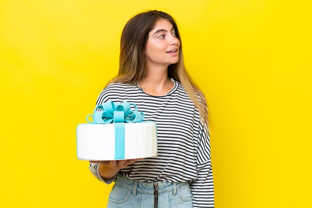 Young caucasian woman holding birthday cake isolated on yellow background looking to the side and smiling