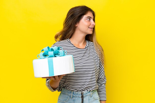 Young caucasian woman holding birthday cake isolated on yellow background looking side