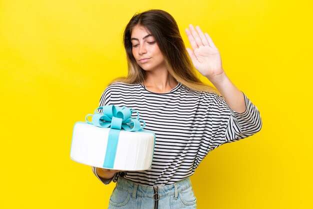 Young caucasian woman holding birthday cake isolated on yellow background making stop gesture and disappointed