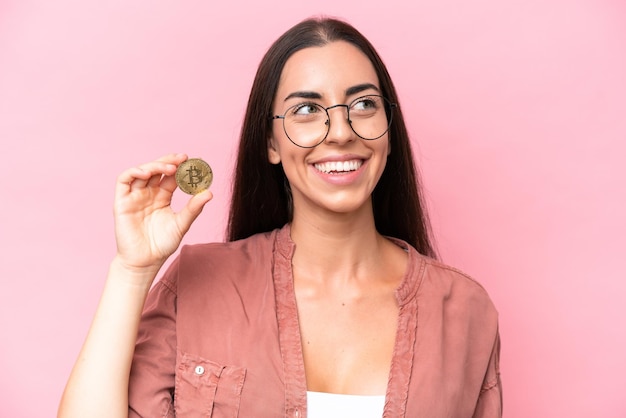 Young caucasian woman holding a Bitcoin isolated on pink background looking up while smiling