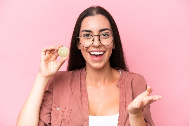 Young caucasian woman holding a Bitcoin isolated on pink background with shocked facial expression