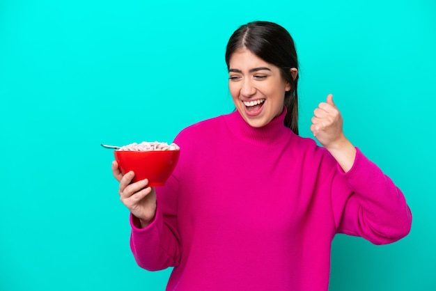 Young caucasian woman holding a bowl of cereals isolated on blue background celebrating a victory