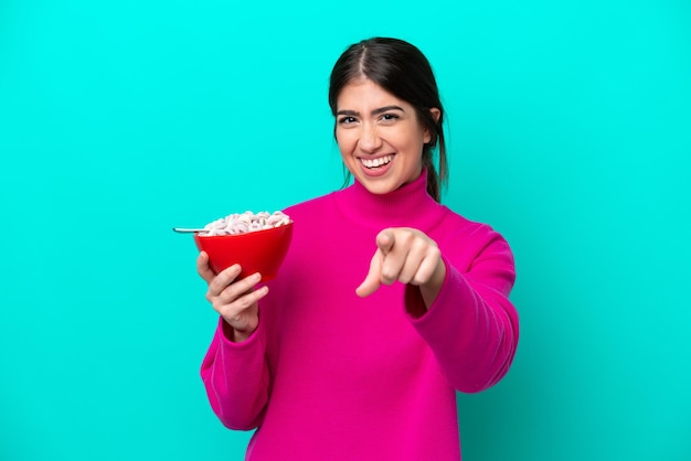 Young caucasian woman holding a bowl of cereals isolated on blue background points finger at you with a confident expression