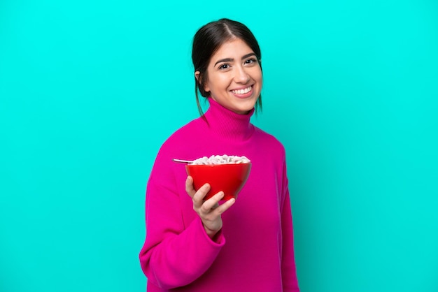 Giovane donna caucasica in possesso di una ciotola di cereali isolata su sfondo blu sorridente molto
