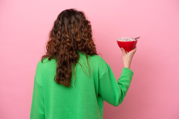 Young caucasian woman holding a bowl of cereals isolated on pink background in back position