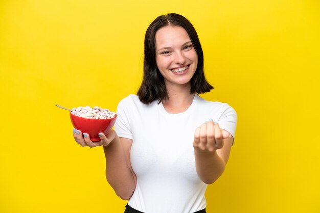 Young caucasian woman holding a bowl of cereals isolated on yellow background inviting to come with hand happy that you came