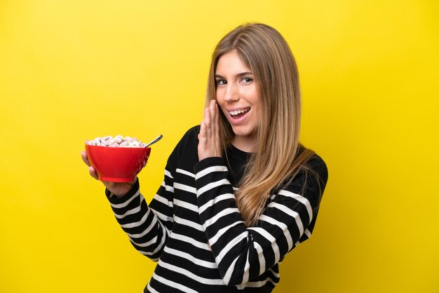 Young caucasian woman holding a bowl of cereals isolated on yellow background whispering something