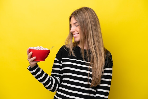 Young caucasian woman holding a bowl of cereals isolated on yellow background with happy expression