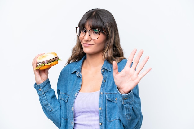Giovane donna caucasica che tiene un hamburger isolato su sfondo bianco che saluta con la mano con espressione felice