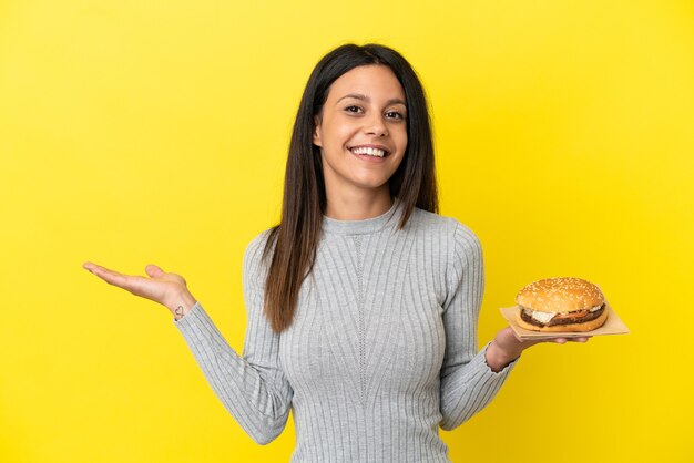 黄色の背景に分離されたハンバーガーを持っている若い白人女性は、来て招待するために手を横に伸ばします