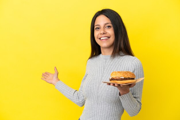 Молодая кавказская женщина держит гамбургер на желтом фоне, протягивая руки в сторону, приглашая прийти