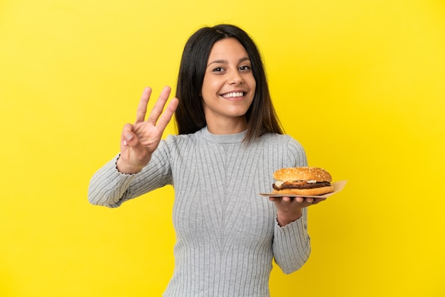 Young caucasian woman holding a burger isolated on yellow background happy and counting three with fingers