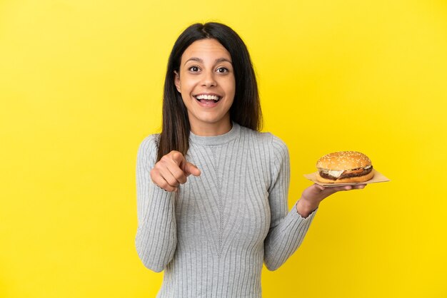 黄色の背景に分離されたハンバーガーを保持している若い白人女性は驚いて正面を指しています