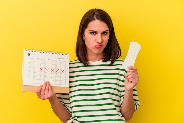 Young caucasian woman holding calendar and compress isolated on yellow background