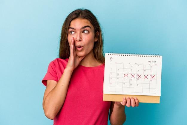 Photo young caucasian woman holding a calendar isolated on pink background is saying a secret hot braking news and looking aside