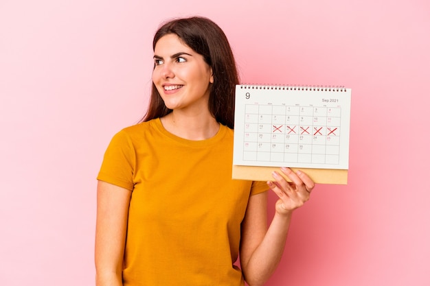 Young caucasian woman holding calendar isolated on pink background looks aside smiling, cheerful and pleasant.